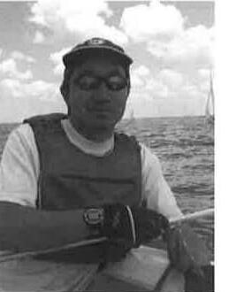 A sailor in a cap looking toward the camera aboard a boat, with another sail visible on the water behind