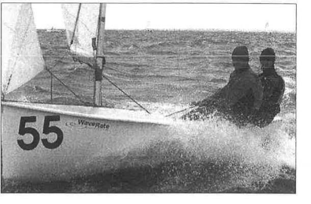 An Albacore, sail number 55, planing hard with spray flying and two crew hiking out