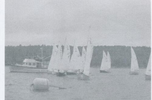 A fleet of Albacores sailing near a small motor cruiser and a mooring buoy
