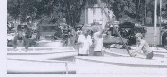 A group of sailors lowering a mast on a trailered Albacore in a boat-park lot