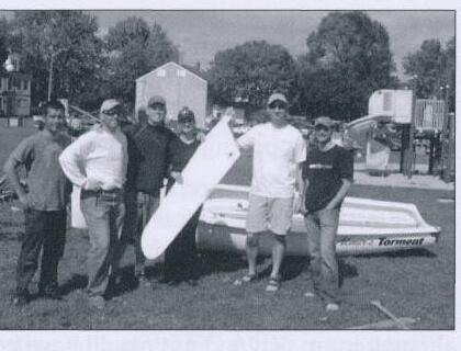 A group of six sailors posing with a spare centreboard on a lawn beside an upturned dinghy