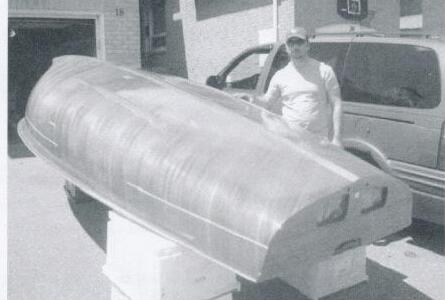 A man standing beside an unpainted wooden Albacore hull upside down on stands in a driveway