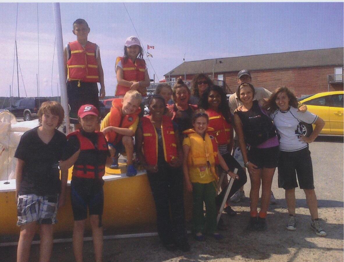 A group of teens and instructors in life jackets posing beside a yellow Albacore hull after a Friday afternoon community sailing school session