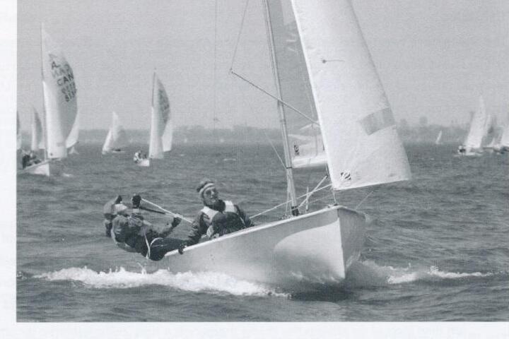 Two women racing an Albacore hard on the rail with a fleet of boats behind, city skyline in the distance