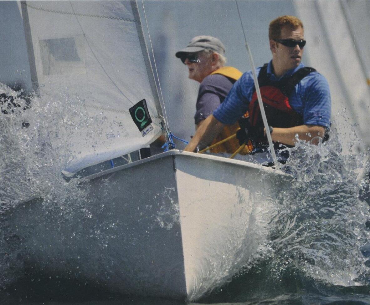 Two sailors on an Albacore crashing through a wave, spray covering the bow