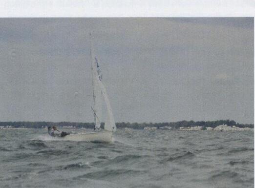 A single Albacore sailing on a grey day with a shoreline in the background
