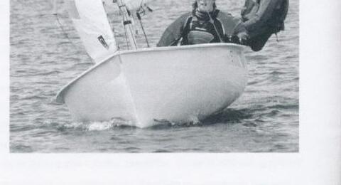 Two sailors racing an Albacore hard, bow-on to the camera, with spray coming off the hull
