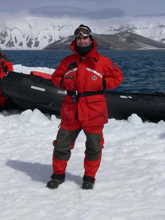 Jeff Beitz in a red survival suit standing on ice in front of a Zodiac, with snow-covered mountains behind