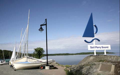 A dockside sailboat mast sculpture with a Sail Parry Sound sign