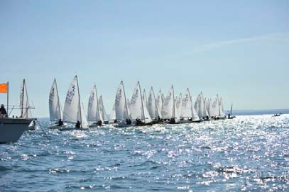 A large fleet of dinghies at a starting line, sails backlit by the sun
