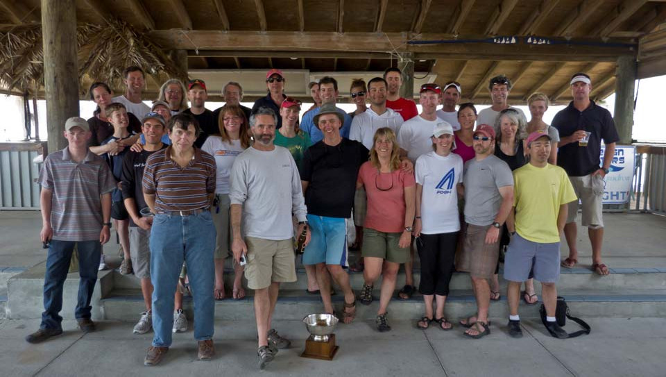 A large group of Albacore sailors posing together under a pavilion at the Mid Winters Regatta in Sarasota, with a trophy in front