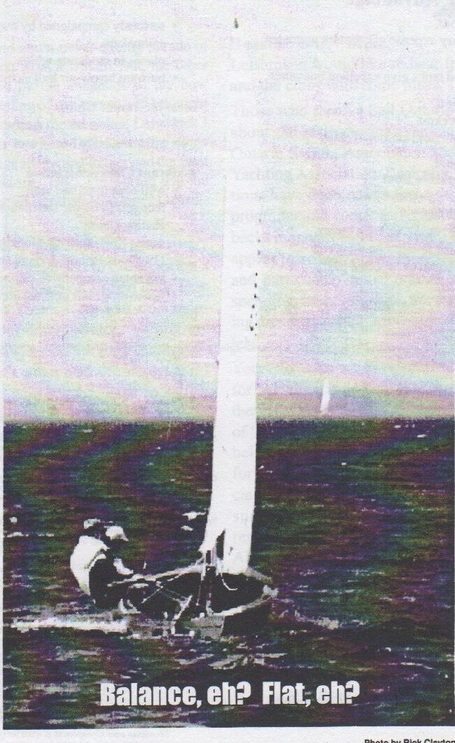 An Albacore sailing upwind on dark water with both crew hiked out, captioned “Balance, eh? Flat, eh?”
