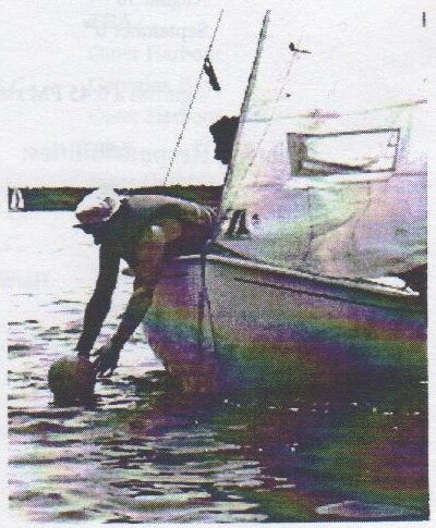 A sailor leaning far out over the bow of an Albacore to grab a mark buoy by hand