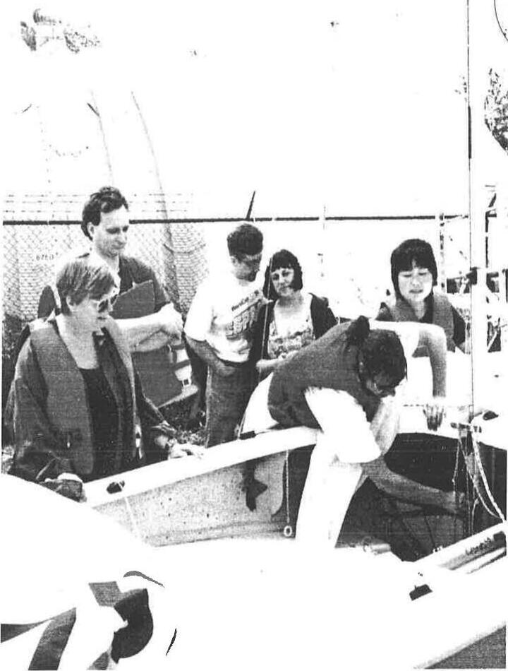 A group of new sailors in life jackets gathered around an Albacore dinghy on shore, one leaning into the boat to rig it