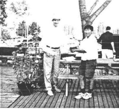 Ian Aukema presenting the first place award to Gord Chu on a wooden deck under a tree
