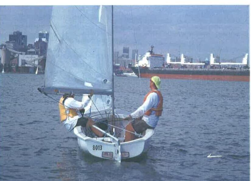 Two sailors on an Albacore dinghy, sail number 8013, with a freighter and city skyline behind them