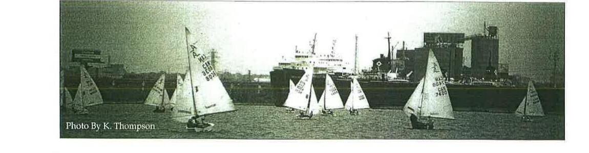 A fleet of Albacores racing on open water with an industrial waterfront and ship in the background
