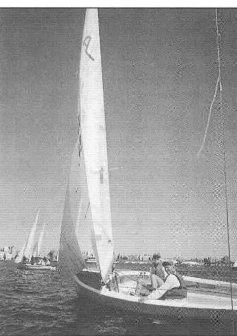 An Albacore with two crew heeling in a breeze, other dinghies and a city skyline in the background