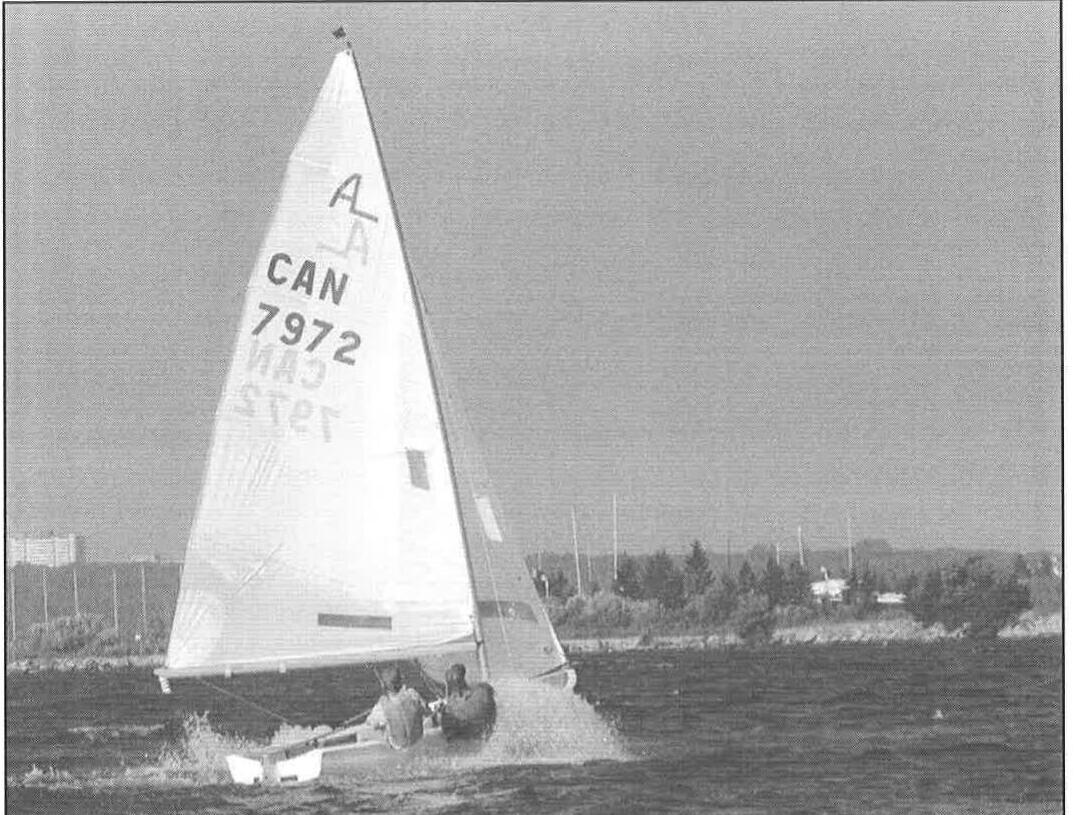 An Albacore planing hard with spray flying, sail CAN 7972, crew hiked out, shoreline behind