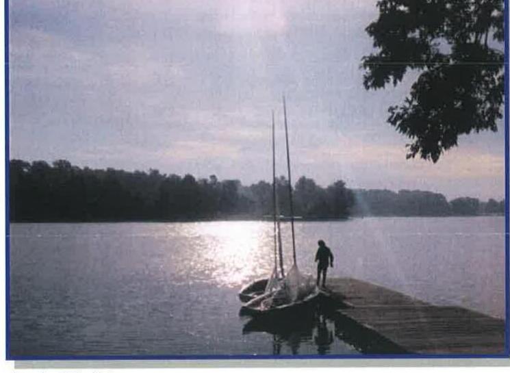 A sailing dinghy and its skipper silhouetted on a dock at sunset on a calm lake