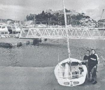 Sailboat and two people on a dock in a Mediterranean harbor town with a hilltop fortress in the background